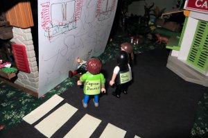 My main character (the brown haired character at the back) and her colleagues are surprised to find the missing man outside their office, lying in a stairwell near the road (the white stripes are a pedestrian crossing). The back wall of the "office" is open (it's a child's play set), so I had to put in a paper wall to ever so slightly increase the realism.